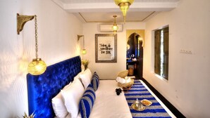 A bedroom with a blue upholstered headboard, a bed with white linens, a decorative wall hanging, and a hallway with a patterned rug leading to a door. - Riad Mazarin des gourmets (Marrakech)