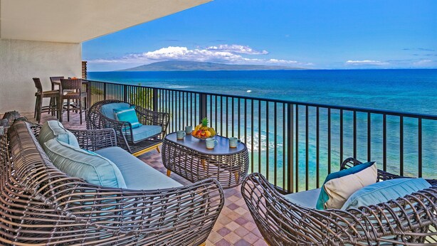 A balcony with wicker furniture, a table with fruit, and a view of the ocean. - Beachfront 2BR condo w stunning ocean views - Kaanapali Shores 702 (Lahaina)