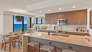A modern kitchen with a large island, wooden cabinets, and a view of the ocean. - Beachfront 2BR condo w stunning ocean views - Kaanapali Shores 702 (Lahaina)