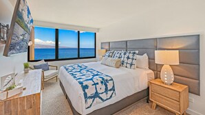 A modern bedroom with a large bed, a bedside table with a lamp, a desk with a chair, and a view of the sea through large windows. - Beachfront 2BR condo w stunning ocean views - Kaanapali Shores 702 (Lahaina)