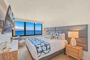 Ein modernes Schlafzimmer mit einem großen Bett, einem Nachttisch mit Lampe, einem Schreibtisch mit Stuhl und Meerblick durch große Fenster.