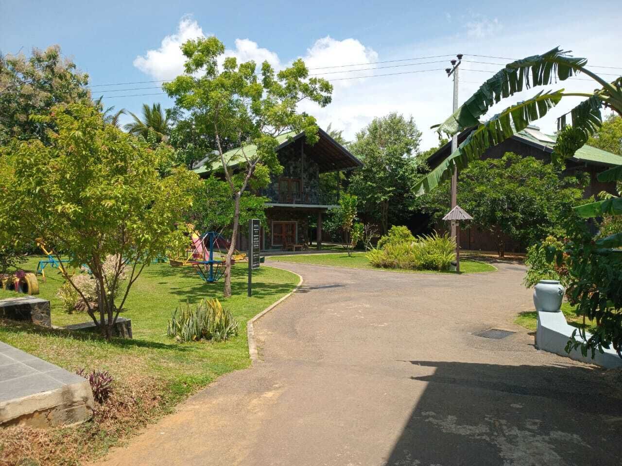 A paved driveway leading to a rustic house with a green roof, surrounded by lush greenery and a small playground.