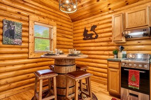 L’intérieur d’une cabane en rondins, avec une table à manger en bois, des tabourets de bar, une cuisine équipée d’une cuisinière et une fenêtre avec des stores.