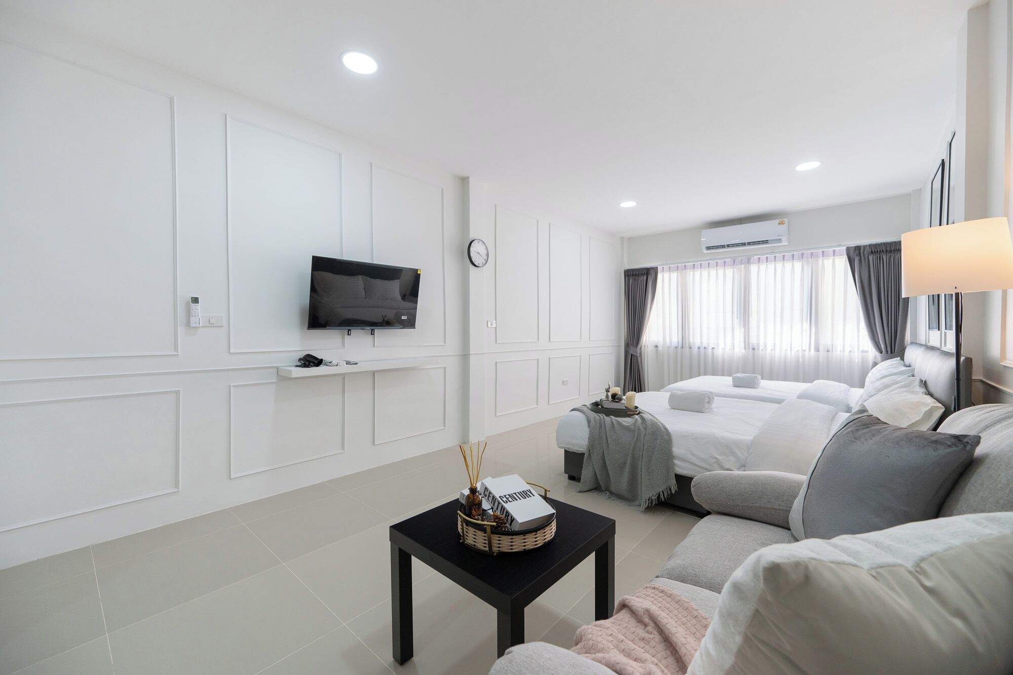 Una camera d'albergo con una TV a schermo piatto montata a parete, un letto con biancheria grigia, un tavolino con un cesto e una lampada.