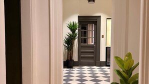 Een gang met een zwart-wit geruit vloerkleed, een kamerplant en een deur met een raam.