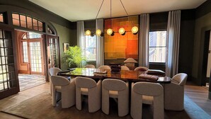 Een eetruimte met een grote houten tafel, zes gestoffeerde stoelen, een hanglamp en uitzicht op buiten door grote ramen.
