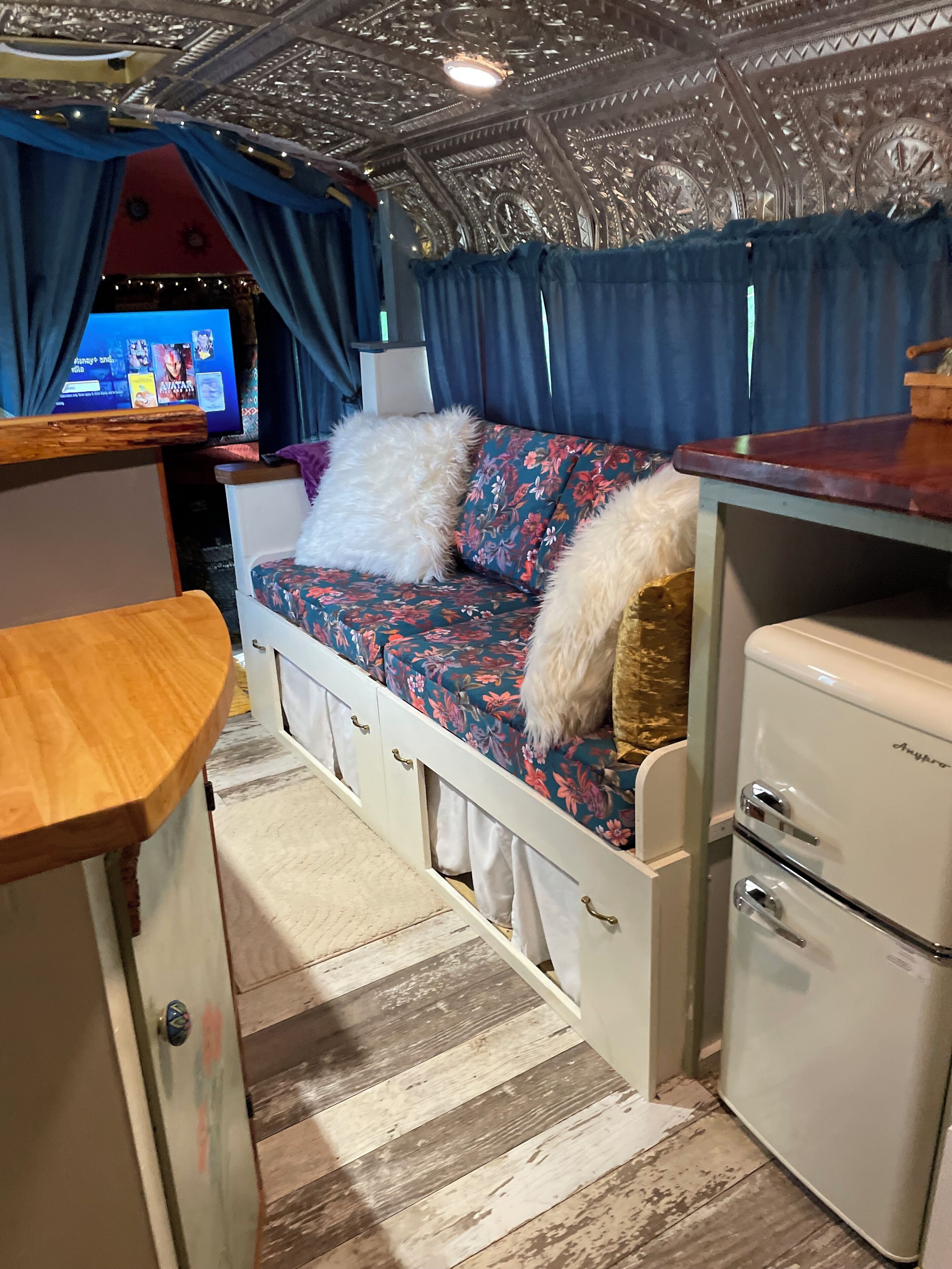 A compact RV interior with a bed, a small kitchenette, and a television.