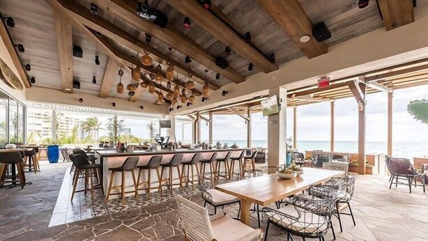 Un amplio espacio exterior para comer con una larga mesa de madera y varias sillas, una barra con taburetes y vista al océano.