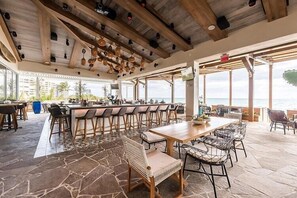 A spacious outdoor dining area with a long wooden table and multiple chairs, a bar with stools, and a view of the ocean. - Casa Palm Serenity | Salt Pool | Close to the Pier (Pompano Beach)