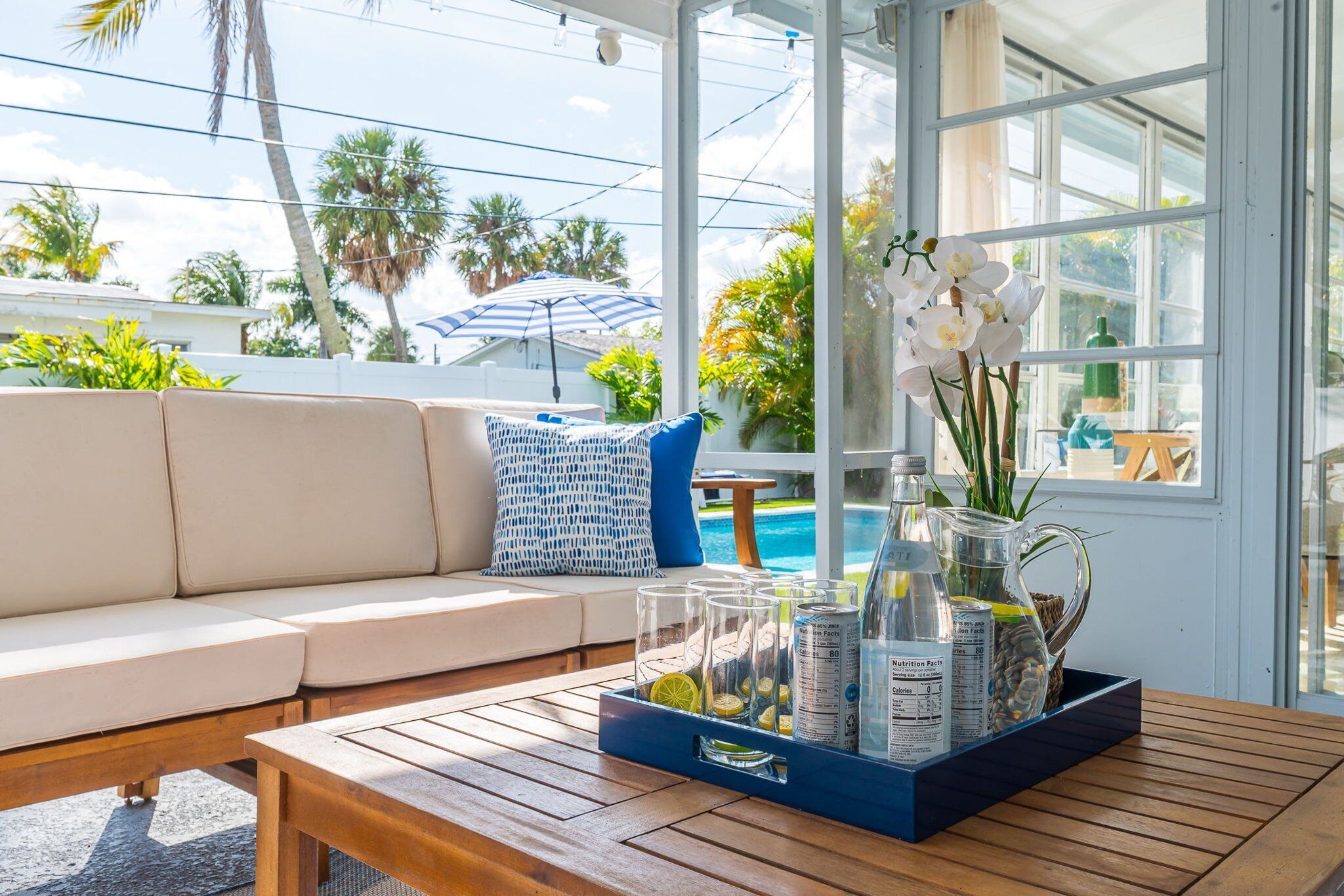 A sunlit patio with a wooden table, a blue tray with drinks and snacks, a white sofa with blue and white cushions, and a view of a swimming pool and palm trees.
