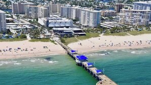 Vista aérea de una zona costera con un muelle largo que se adentra en el agua, rodeado de playa y edificios urbanos.