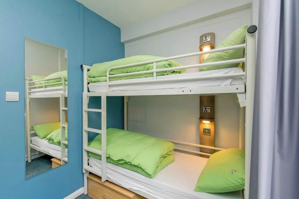 Una habitación con dos literas, cada una con colchón y almohada verdes. Hay un espejo en la pared izquierda y un interruptor de luz encima de la litera superior.