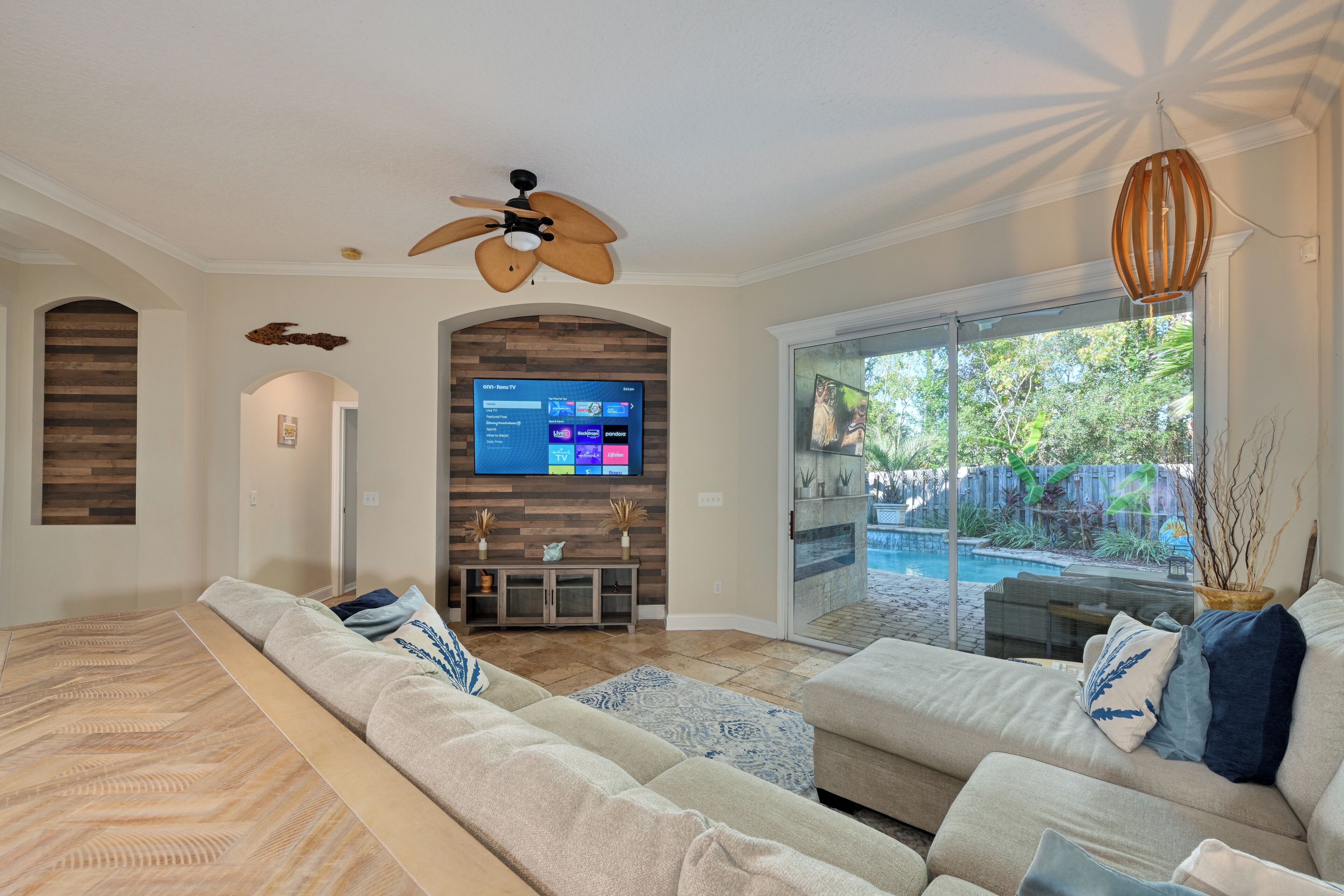 A modern living room with a large sectional sofa, a flat-screen TV mounted on a wooden wall, and a ceiling fan.