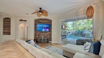 A modern living room with a large sectional sofa, a flat-screen TV mounted on a wooden wall, and a ceiling fan.