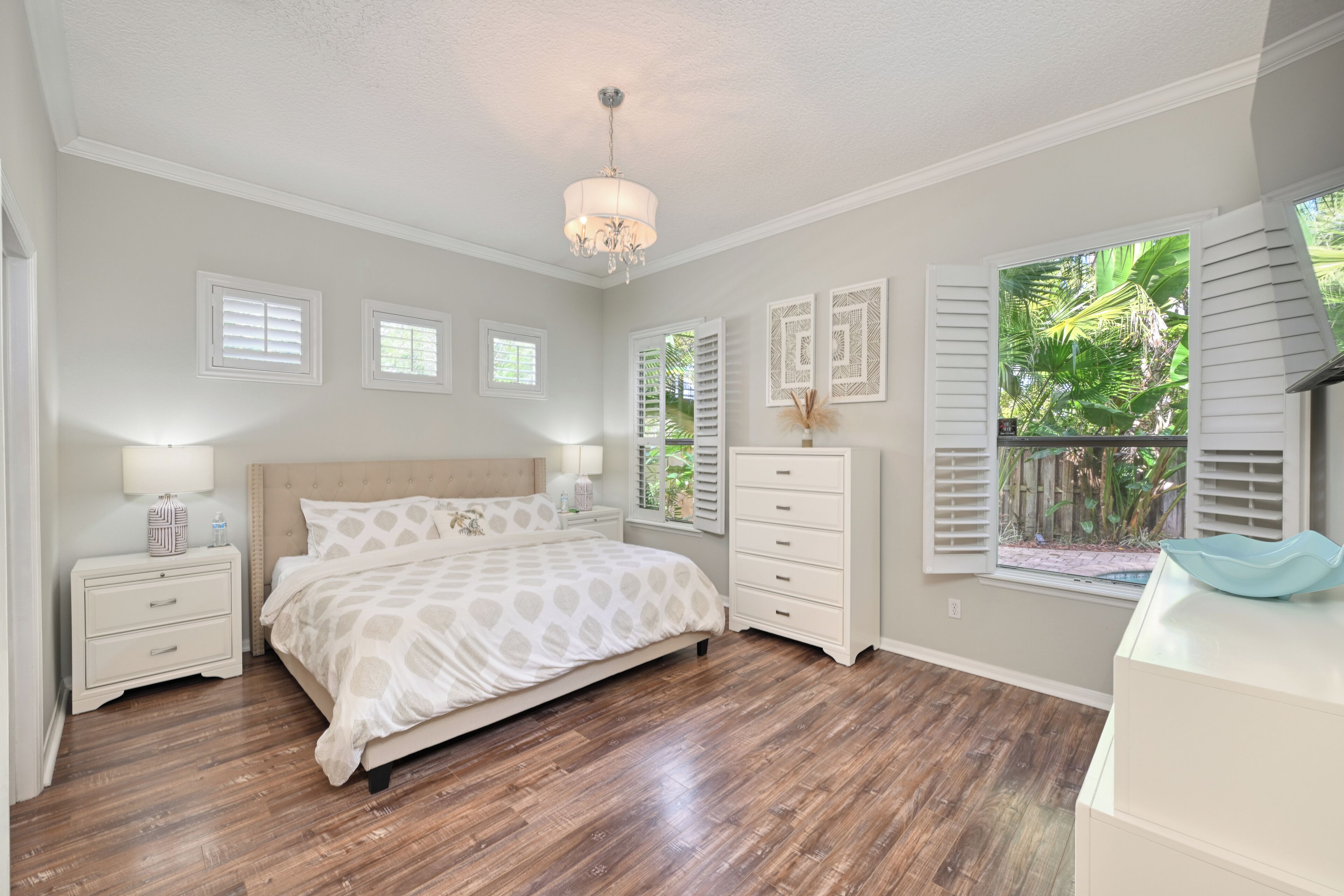 A bedroom with a bed, nightstands, a dresser, a chandelier, and a window with shutters.