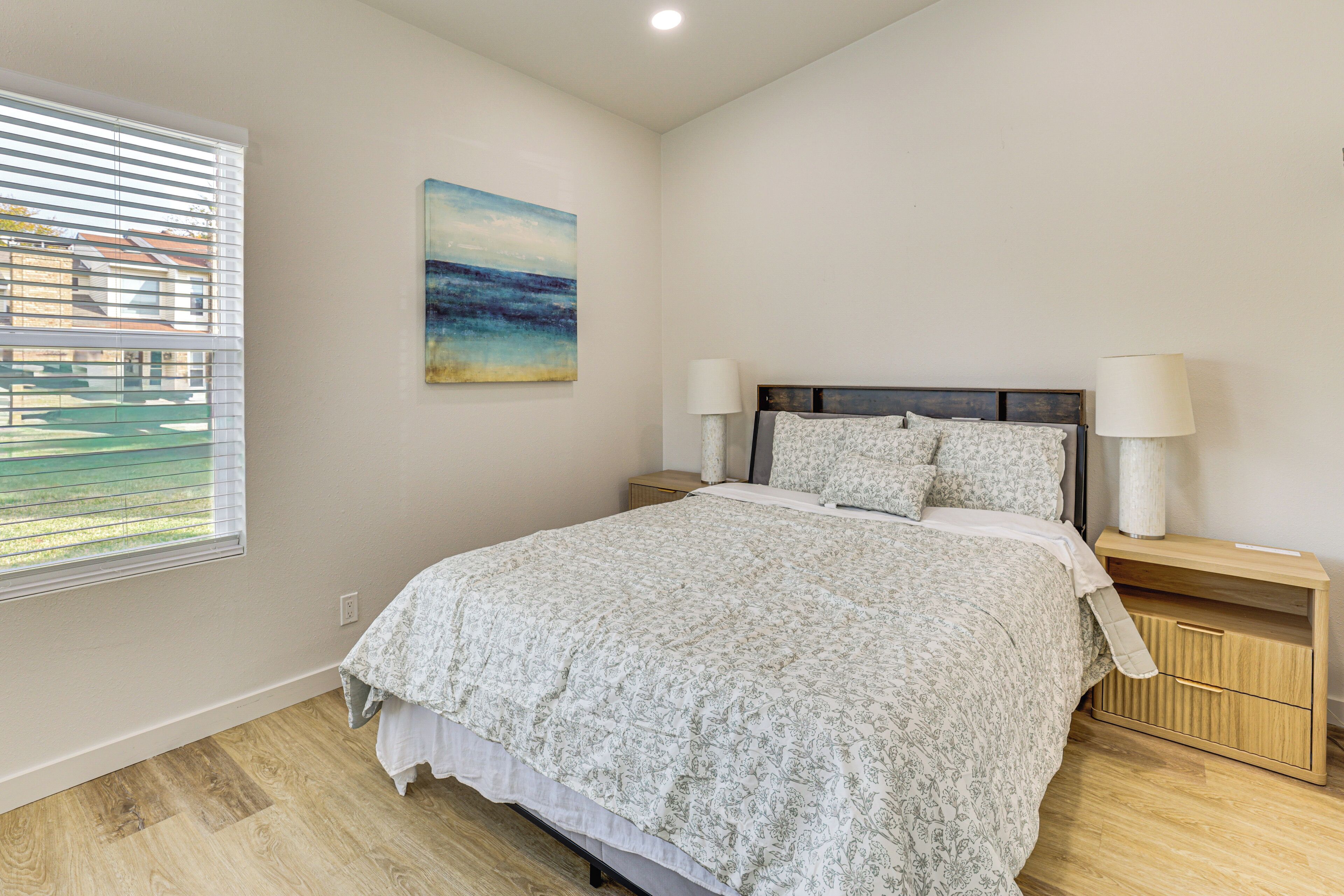 A bedroom with a bed, two bedside tables, a nightstand, a painting, and a window with blinds.