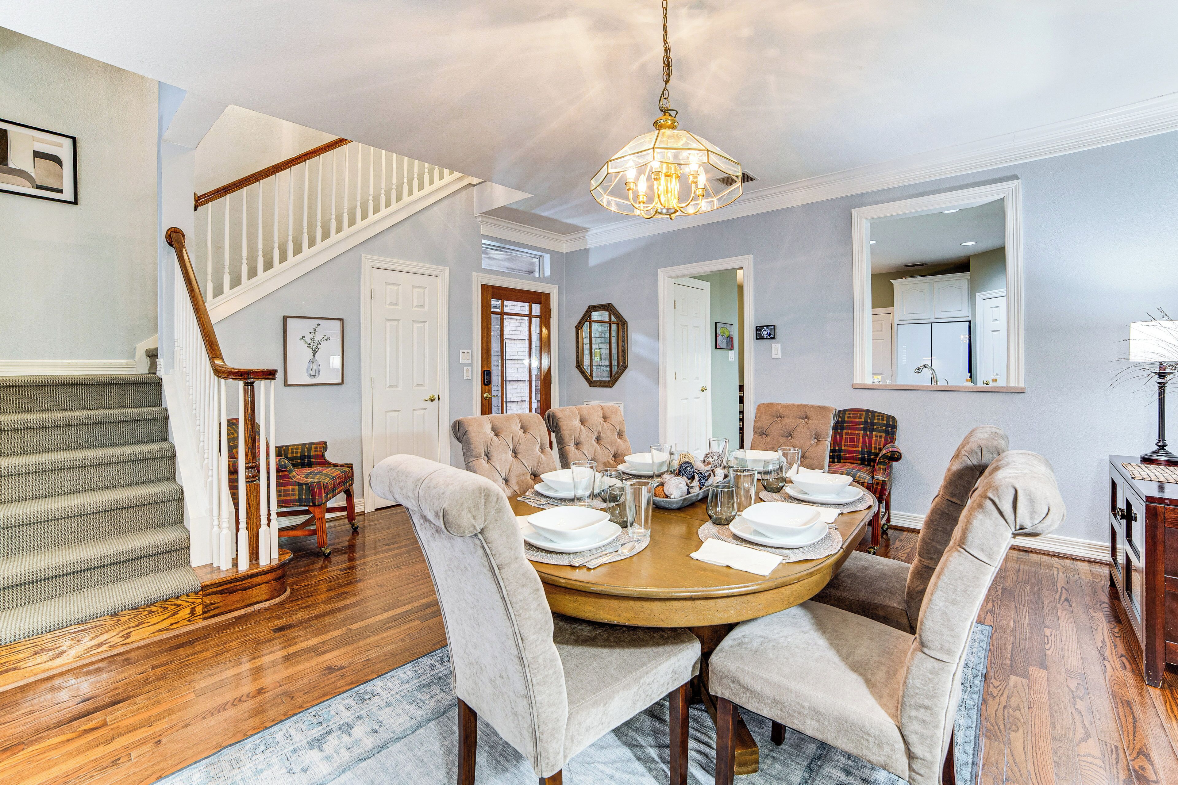 A dining area with a round table set for six, surrounded by upholstered chairs. A staircase with wooden railings is visible to the left.
