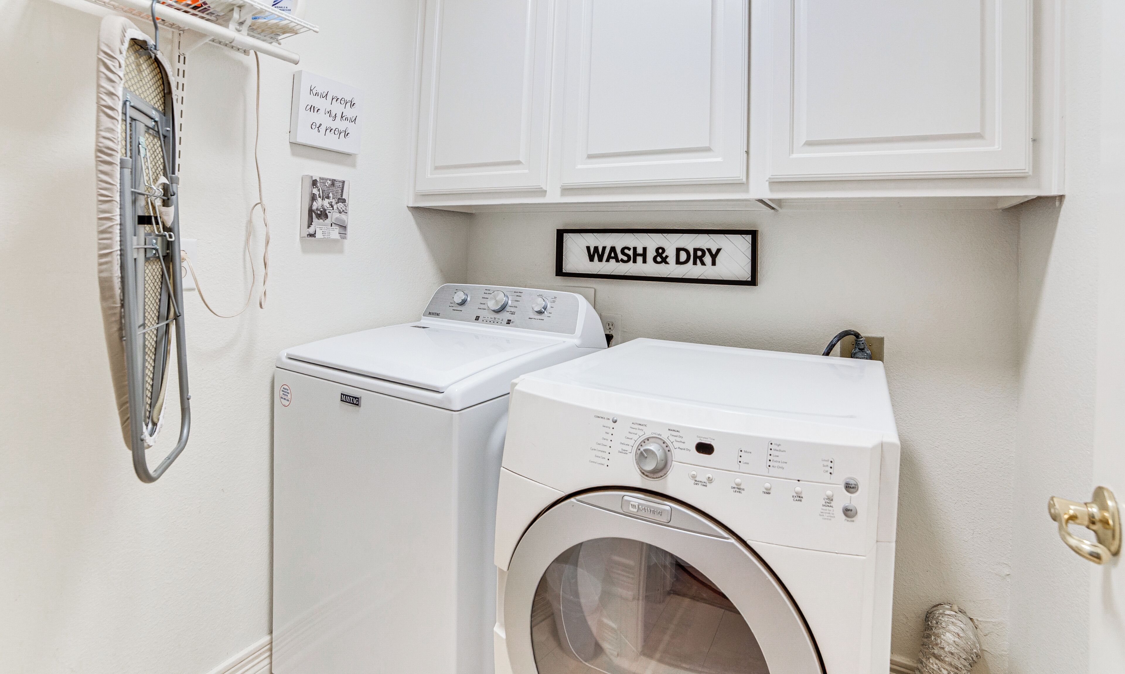 Een wasruimte met een wasmachine en droger, een 'WASH & DRY' bord en een klein plankje met een foto en citaat aan de muur.