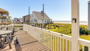 Una terraza de madera con barandal blanco, con vista a una playa y un área verde con una casa al fondo.