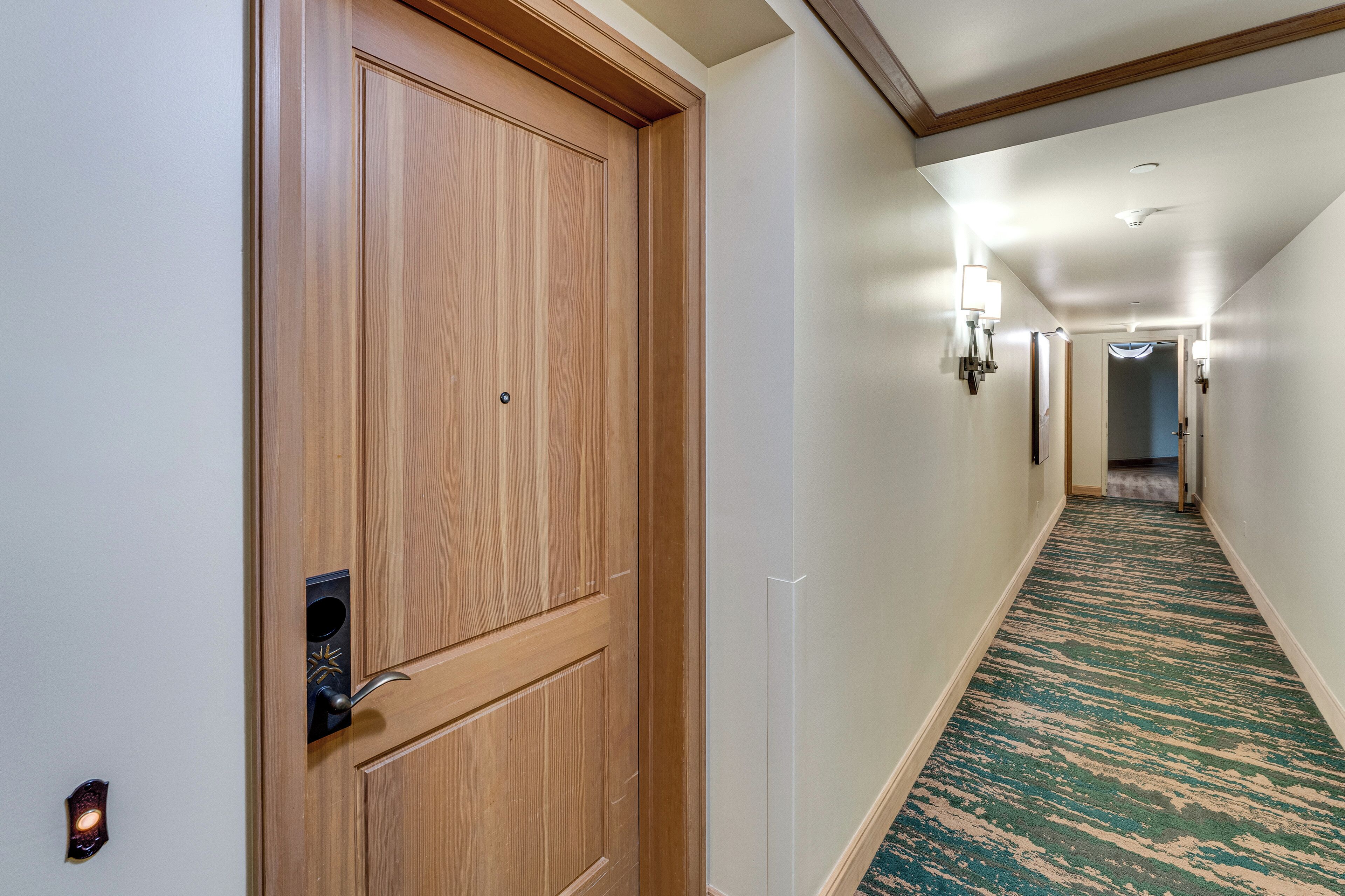 A hotel hallway with a wooden door, a patterned carpet, and wall-mounted lights.