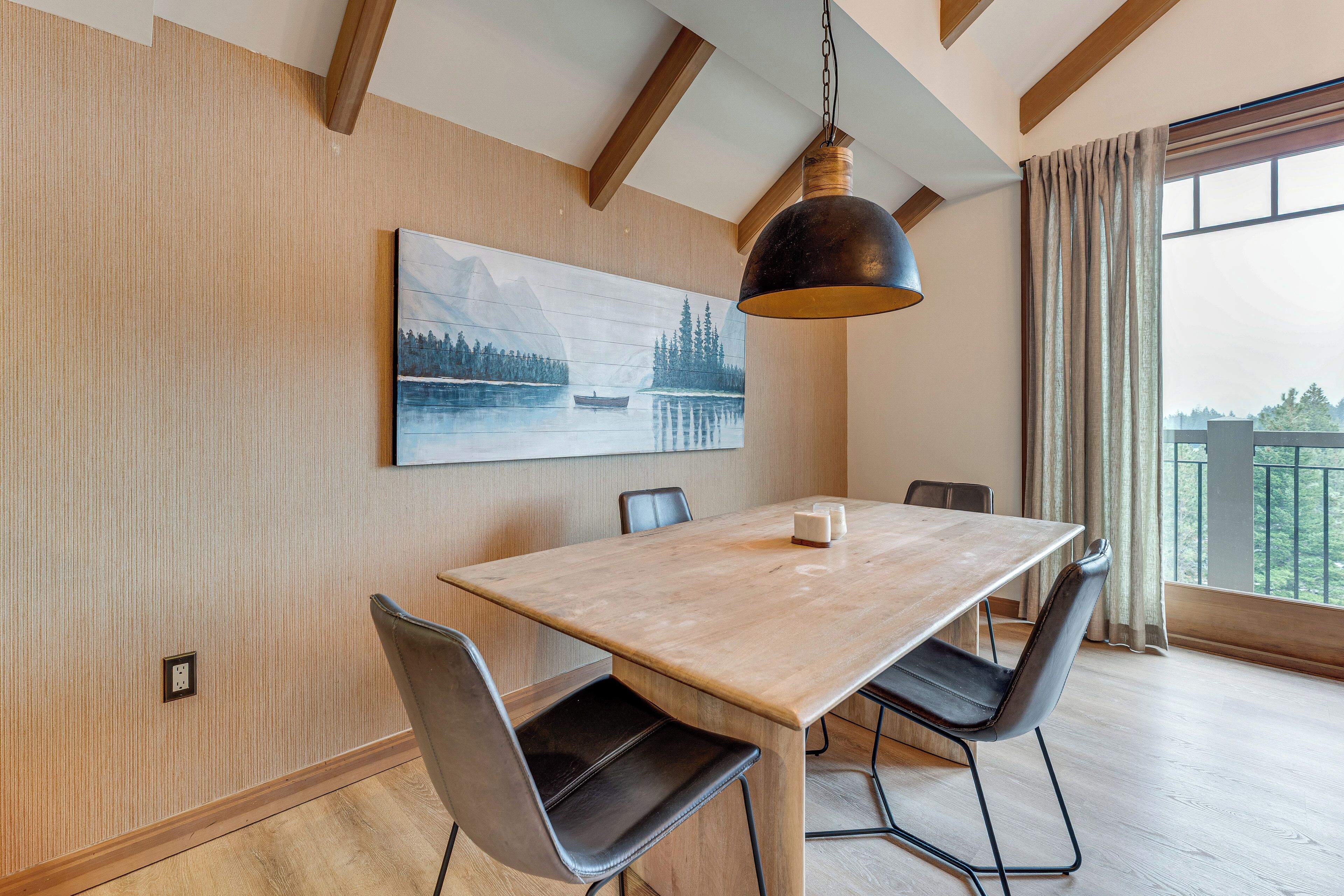 Un coin repas avec une table et des chaises en bois, un tableau représentant un lac et une vue sur la verdure grâce à de grandes fenêtres.