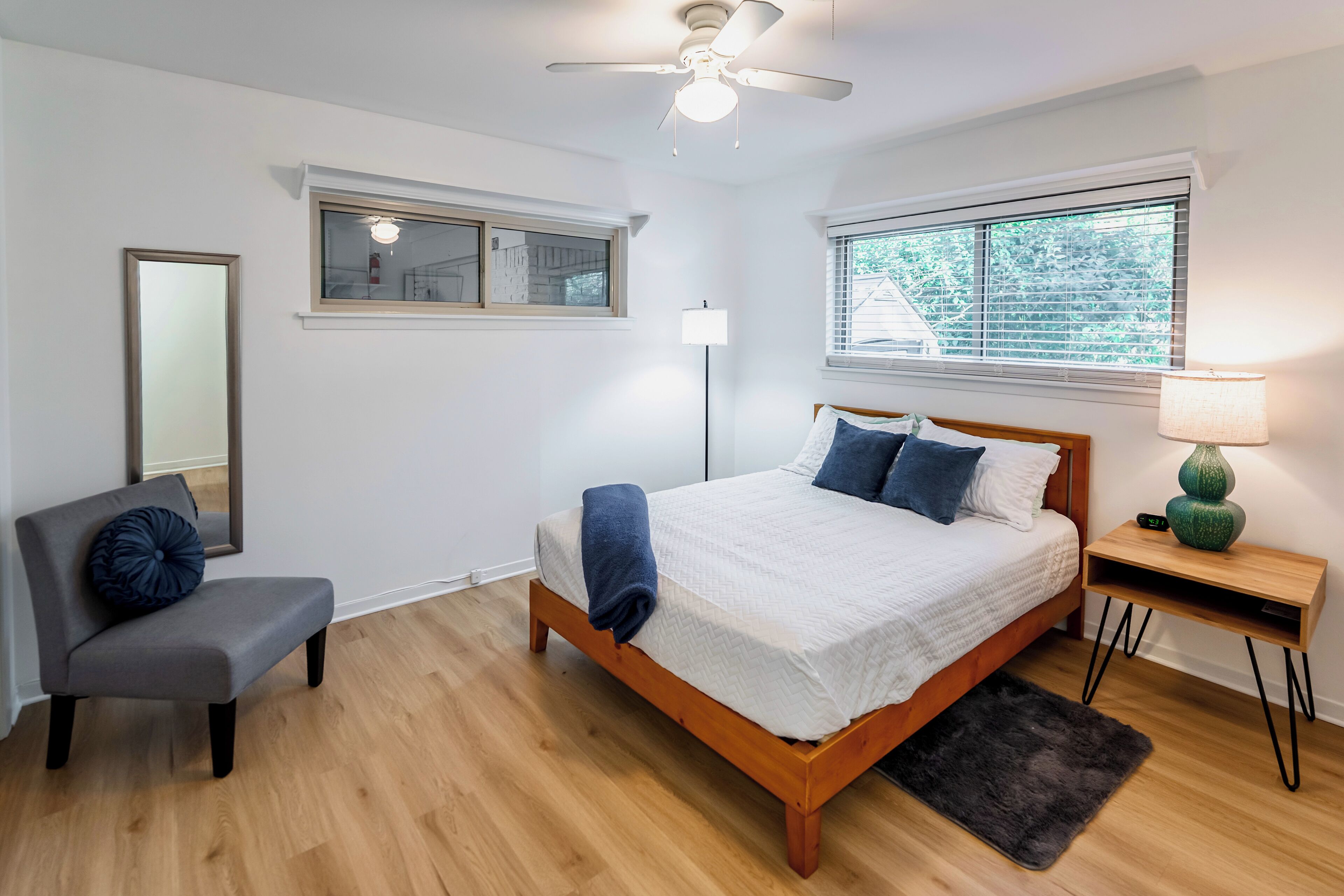A bedroom with a bed, a chair, a bedside table, a lamp, a ceiling fan, a window, and a mirror.