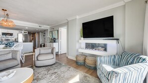 A modern living room with a flat-screen TV, grey sofas, a coffee table, and a kitchen in the background. - Steps to Sand! 8th-floor North Myrtle Beach Condo (North Myrtle Beach)