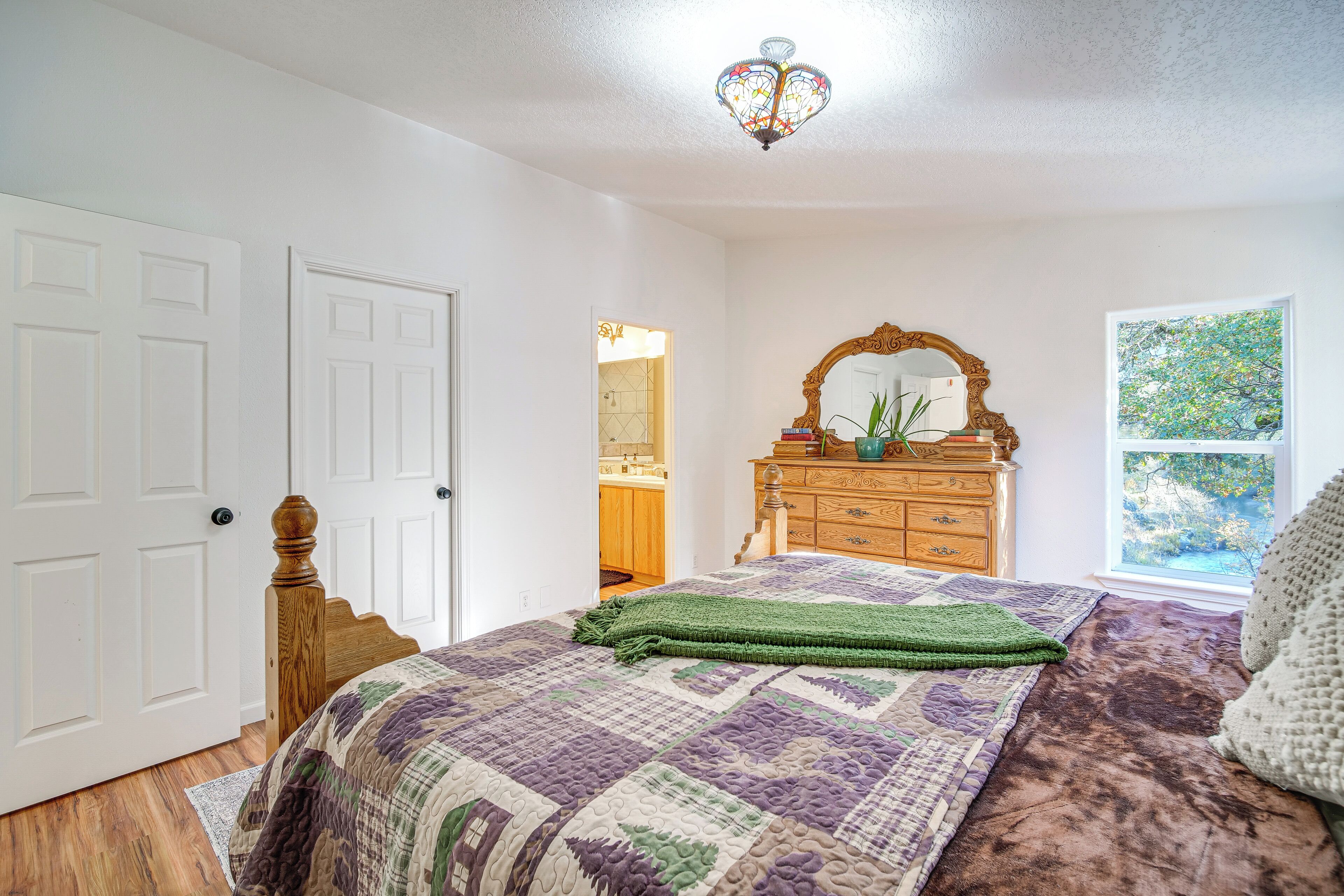 A bedroom with a wooden bed, a green blanket, a wooden dresser, a mirror, and a window with a view of trees.