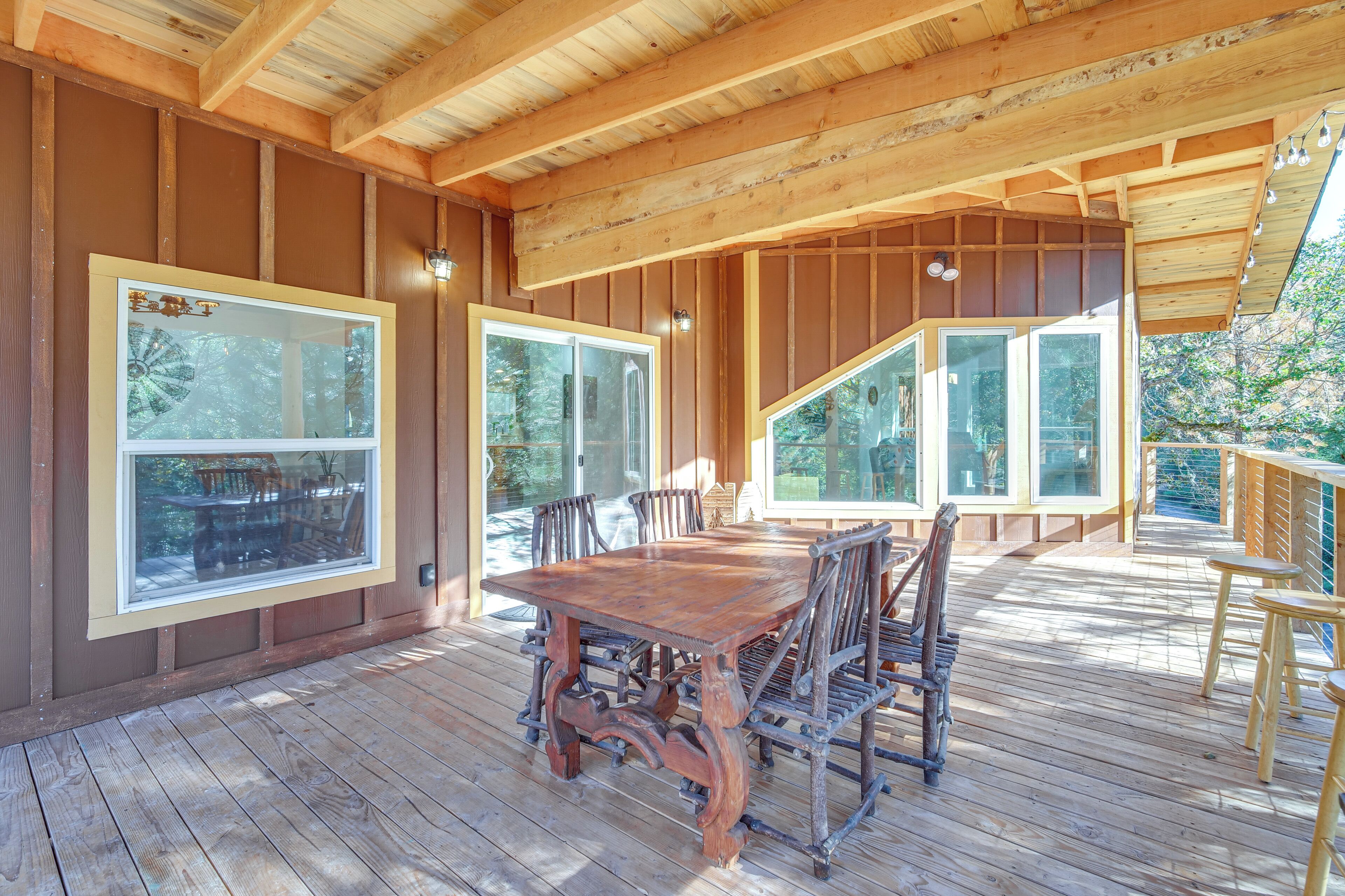 Une terrasse en bois avec une grande table à manger et des chaises, de grandes fenêtres et une balustrade en bois.