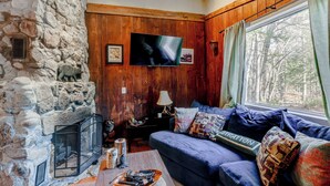 A cozy living room with a stone fireplace, a blue sofa, and a large window with curtains. - 4 Mi to Stratton Mountain Resort! Cabin w/ Hot Tub (Jamaica)