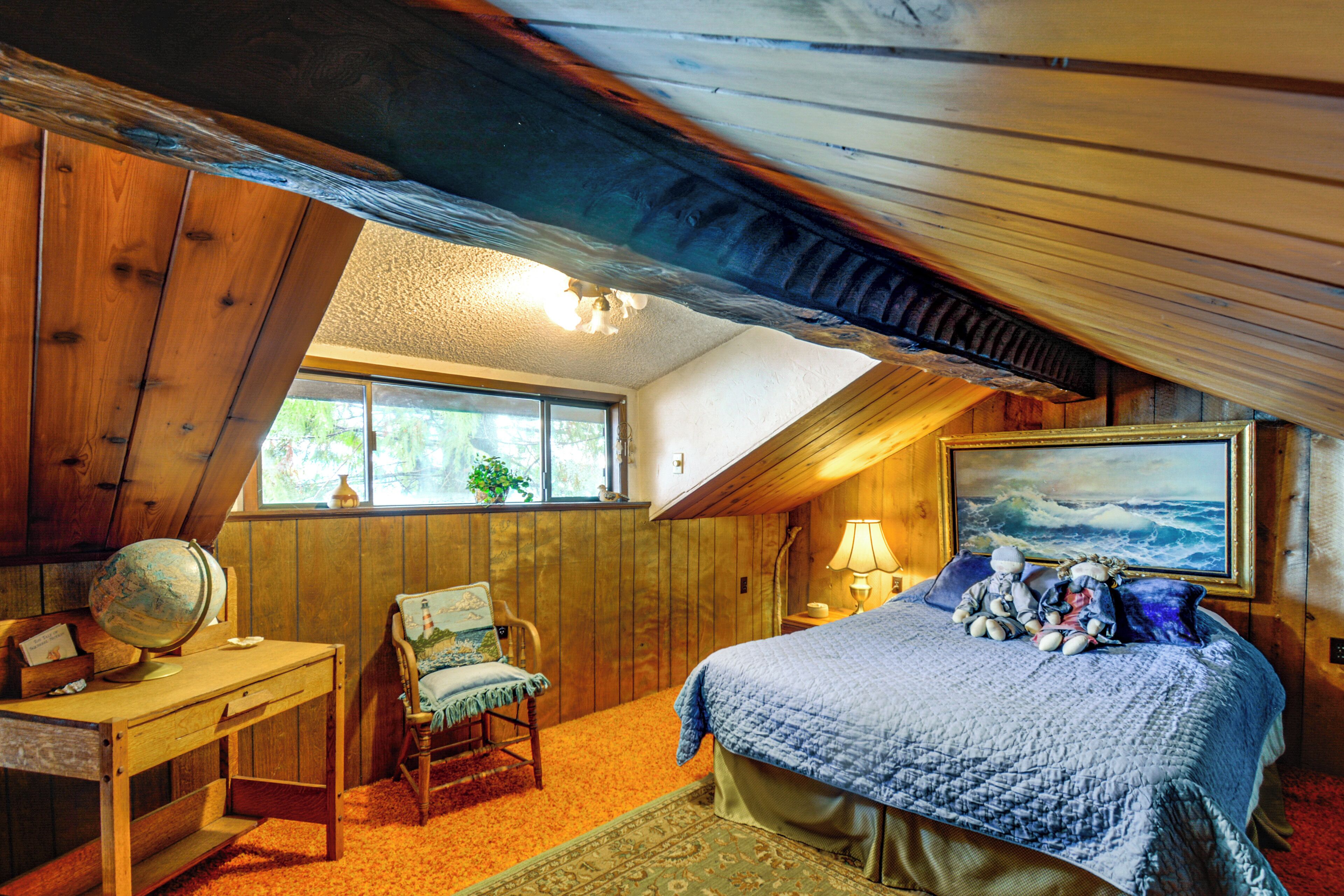 Une chambre mansardée chaleureuse avec un plafond en bois, un bureau surmonté d’un globe terrestre, une chaise ornée d’une peinture marine, un lit avec une courtepointe et des oreillers, et un tableau représentant l’océan.