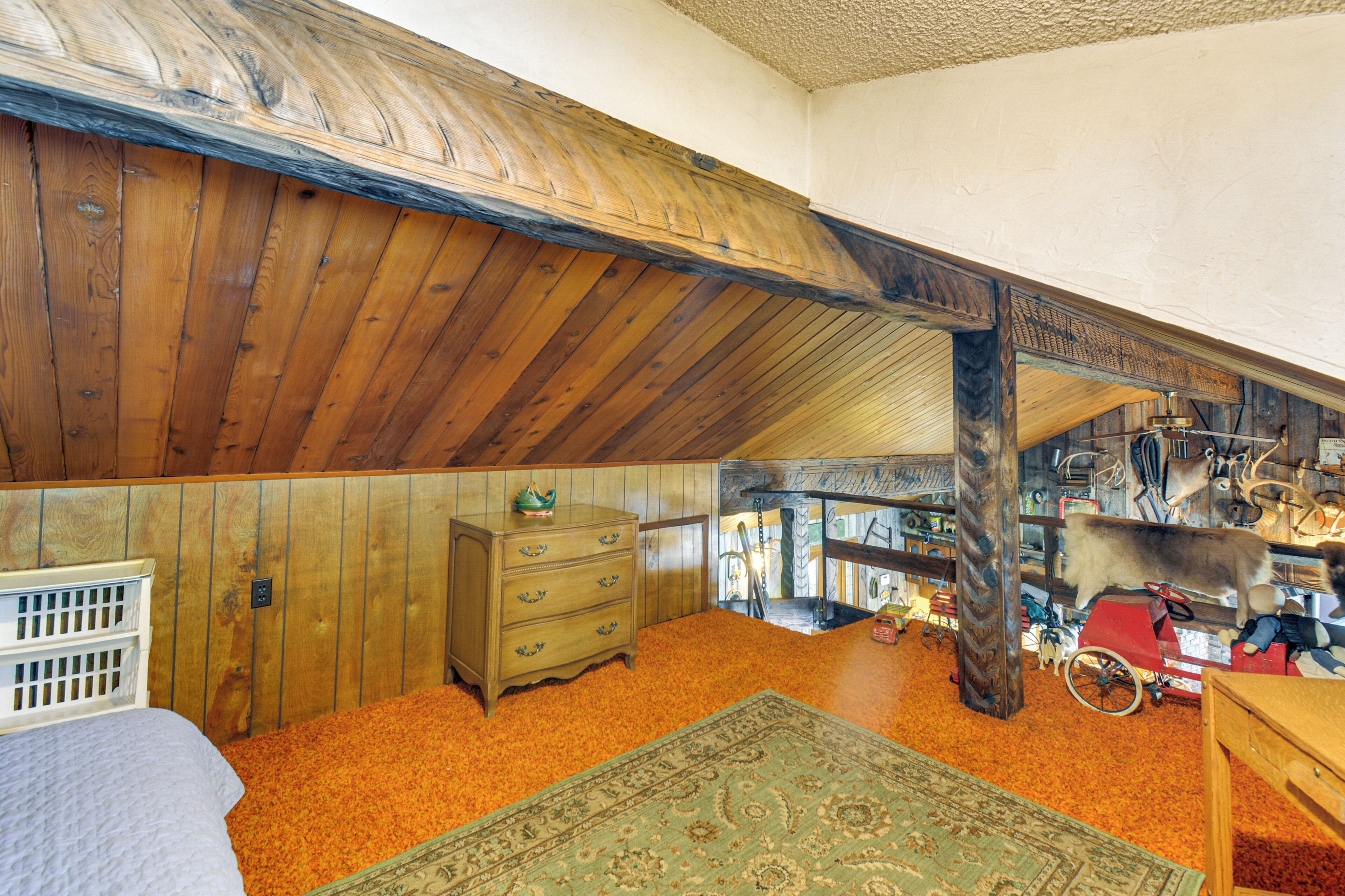 A room with wooden walls and a sloped ceiling, a wooden dresser, a rug, and various items including a toy wagon and stuffed animals.