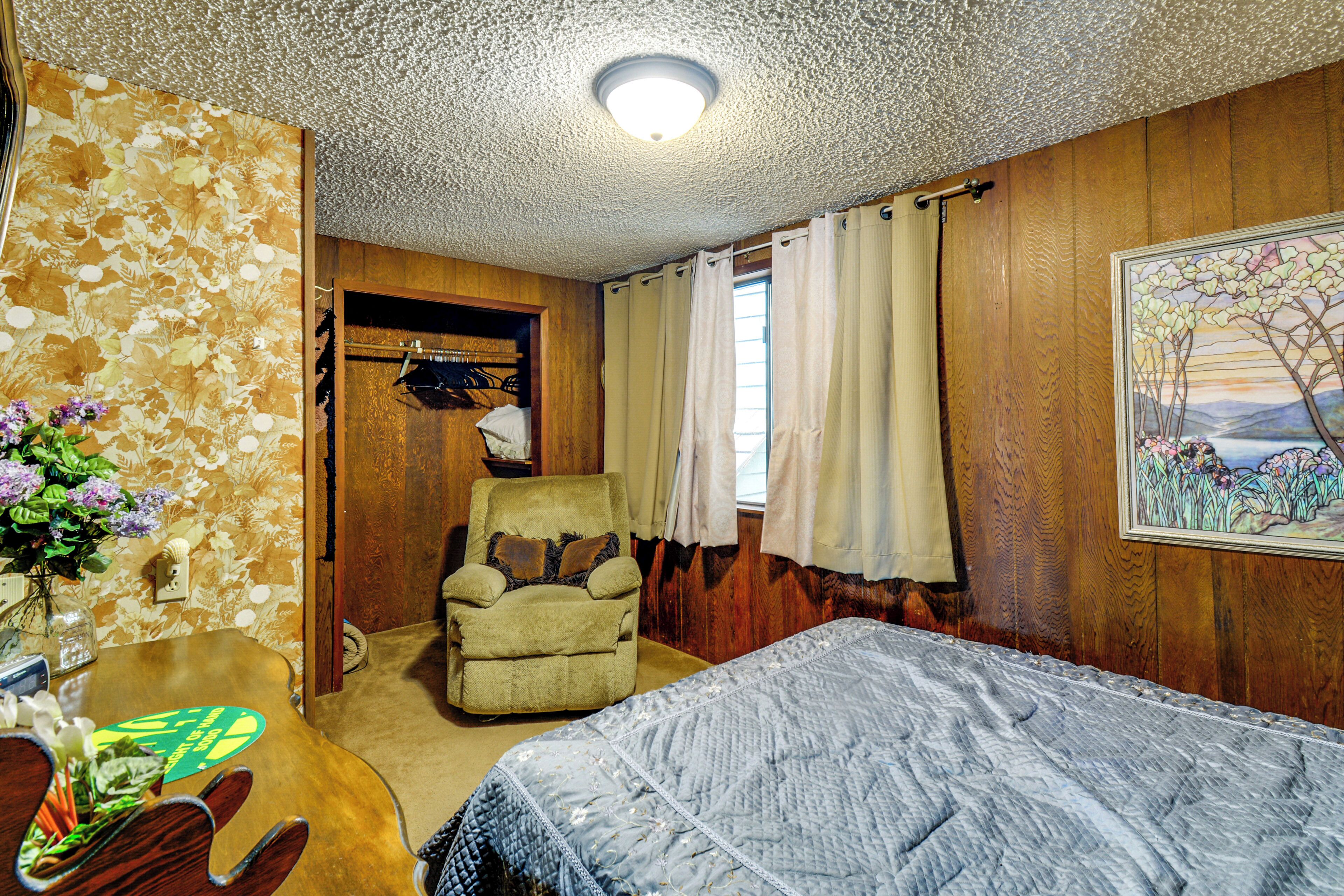 A bedroom with a bed, a chair, a table with a vase of flowers, a coat rack, and a painting on the wall.