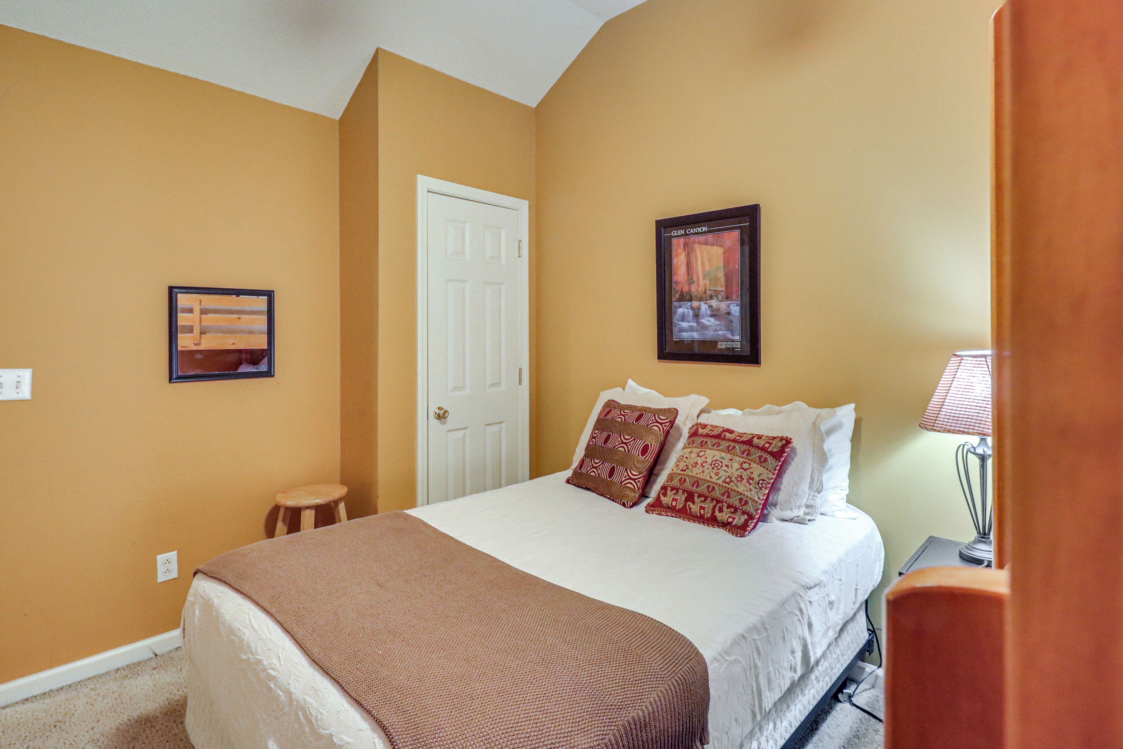A bedroom with a bed, a door, a framed picture, and a nightstand with a lamp.