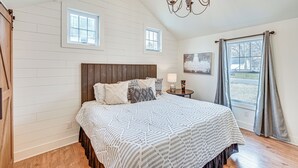 Une chambre à coucher avec un grand lit, une tête de lit en bois, deux fenêtres, un lustre et une vue sur l’extérieur.