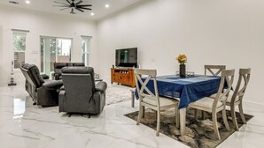 A spacious living room with a dining area, a ceiling fan, a flat-screen TV, and a large window.