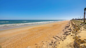A beach with a clear sky, sandy shore, and a rocky cliff with palm trees. - Steps to Shore: Ormond Beach Condo w/ Pool Access! (Ormond Beach)