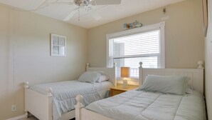A bedroom with two single beds, a ceiling fan, a window with blinds, and a small bedside table with a lamp. - Steps to Shore: Ormond Beach Condo w/ Pool Access! (Ormond Beach)