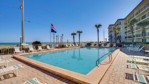 A swimming pool with lounge chairs, palm trees, and a view of the ocean. - Steps to Shore: Ormond Beach Condo w/ Pool Access! (Ormond Beach)
