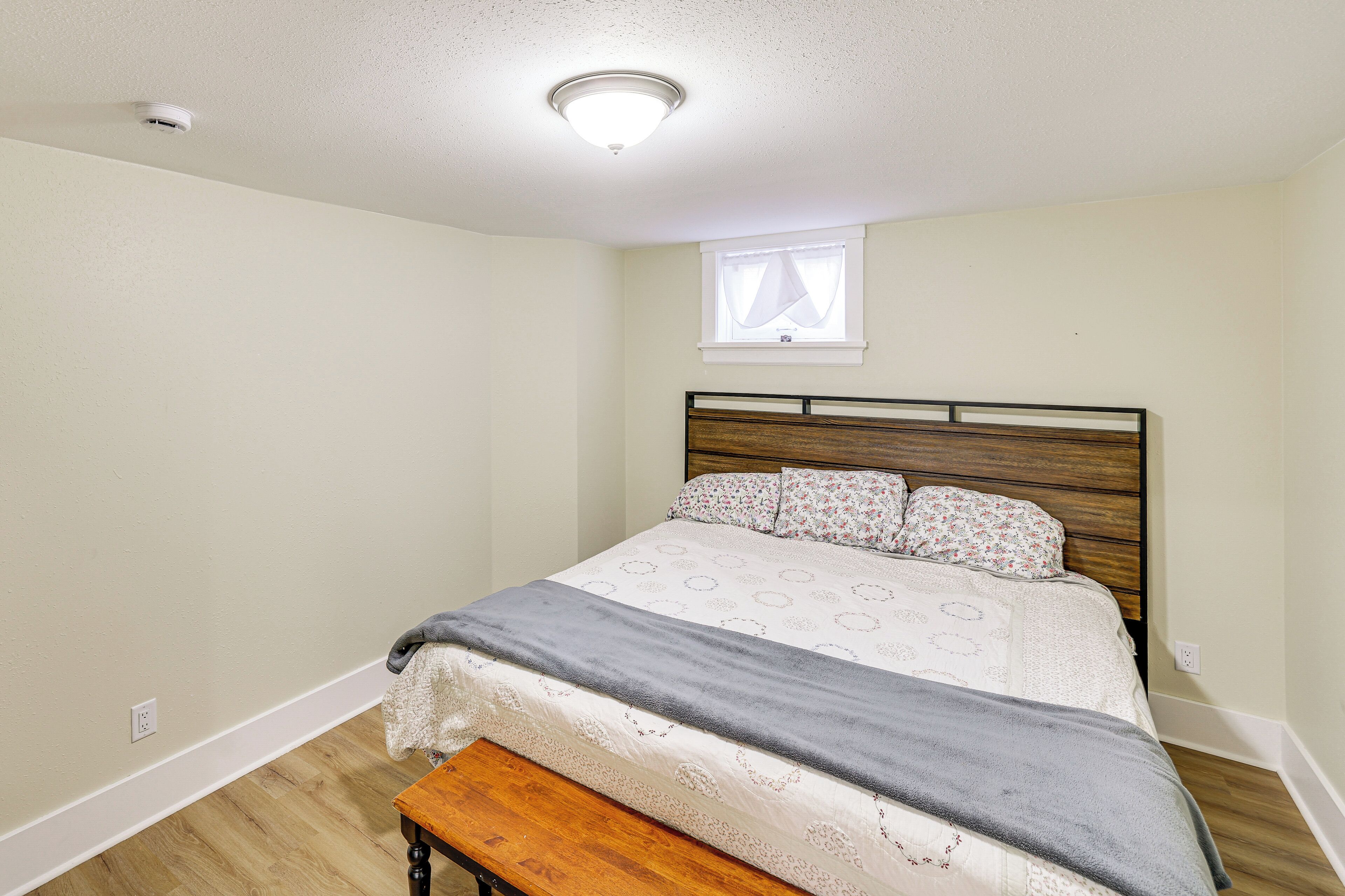A bedroom with a wooden headboard, a bed with floral bedding, a bench at the foot of the bed, and a window with sheer curtains.
