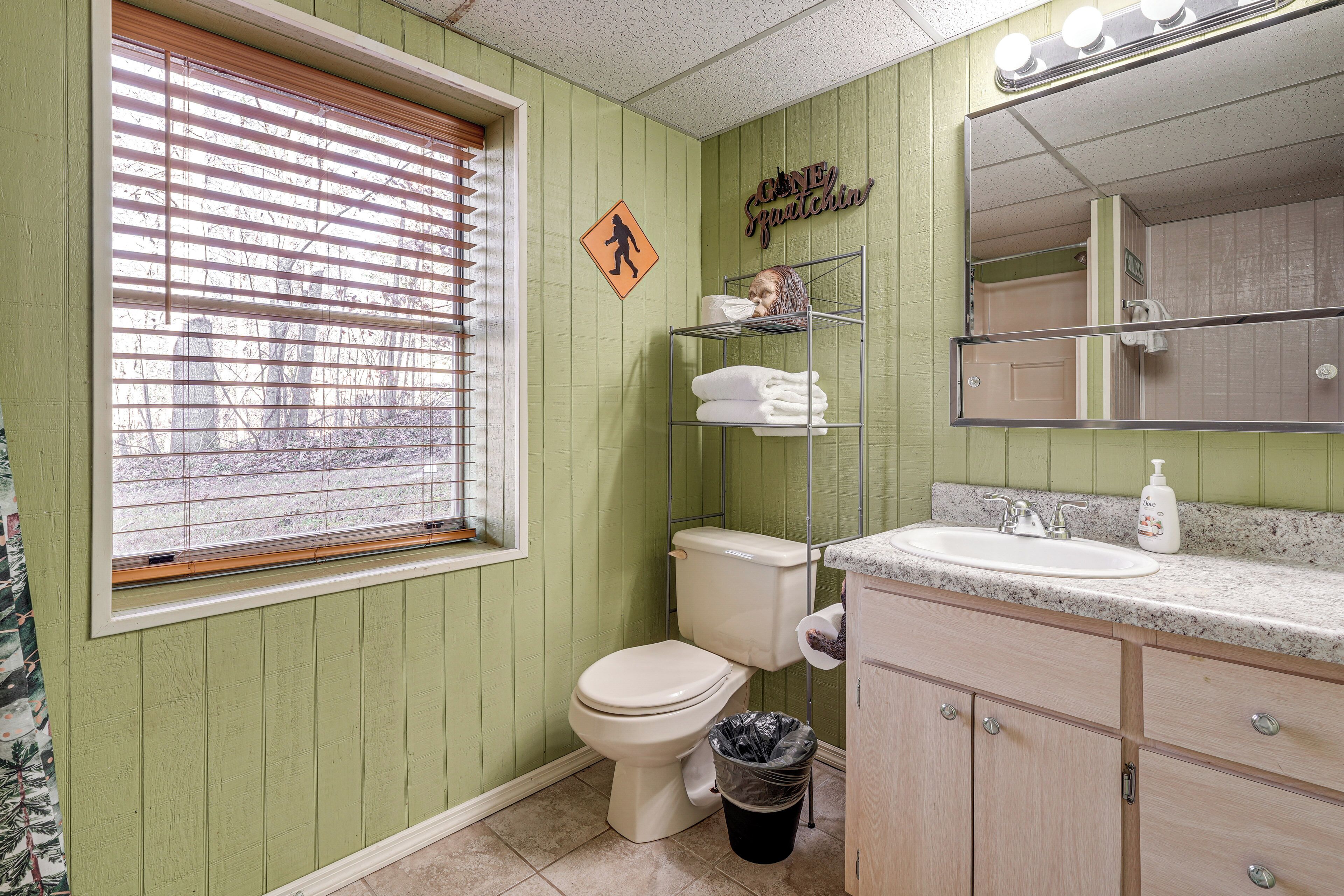 A bathroom with a toilet, sink, and vanity. There is a window with blinds, a shower curtain, and a sign on the wall.