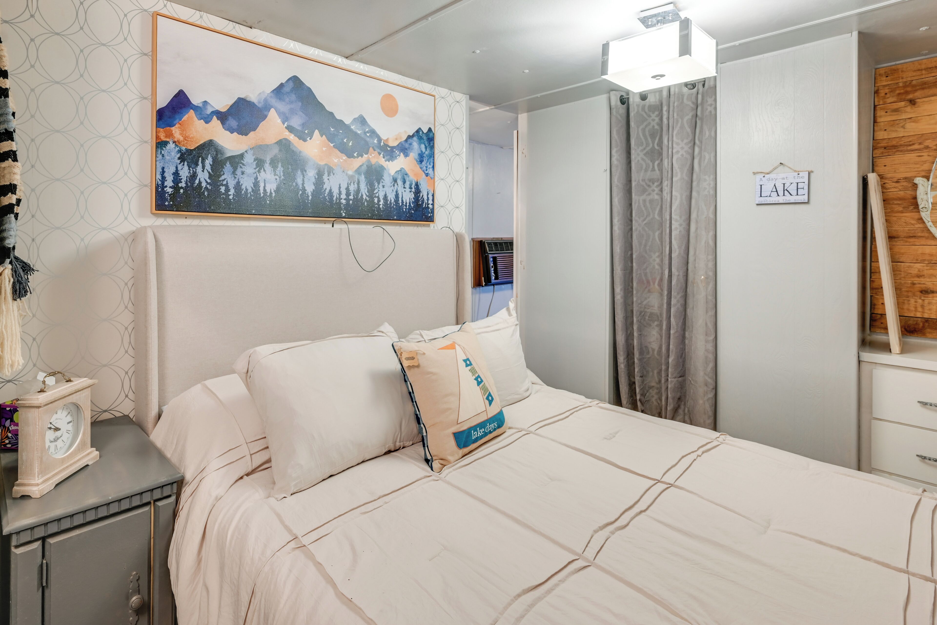 A neatly made bed with a mountain painting above it, a bedside table with a clock, and a sign on the wall that reads "LAKE".