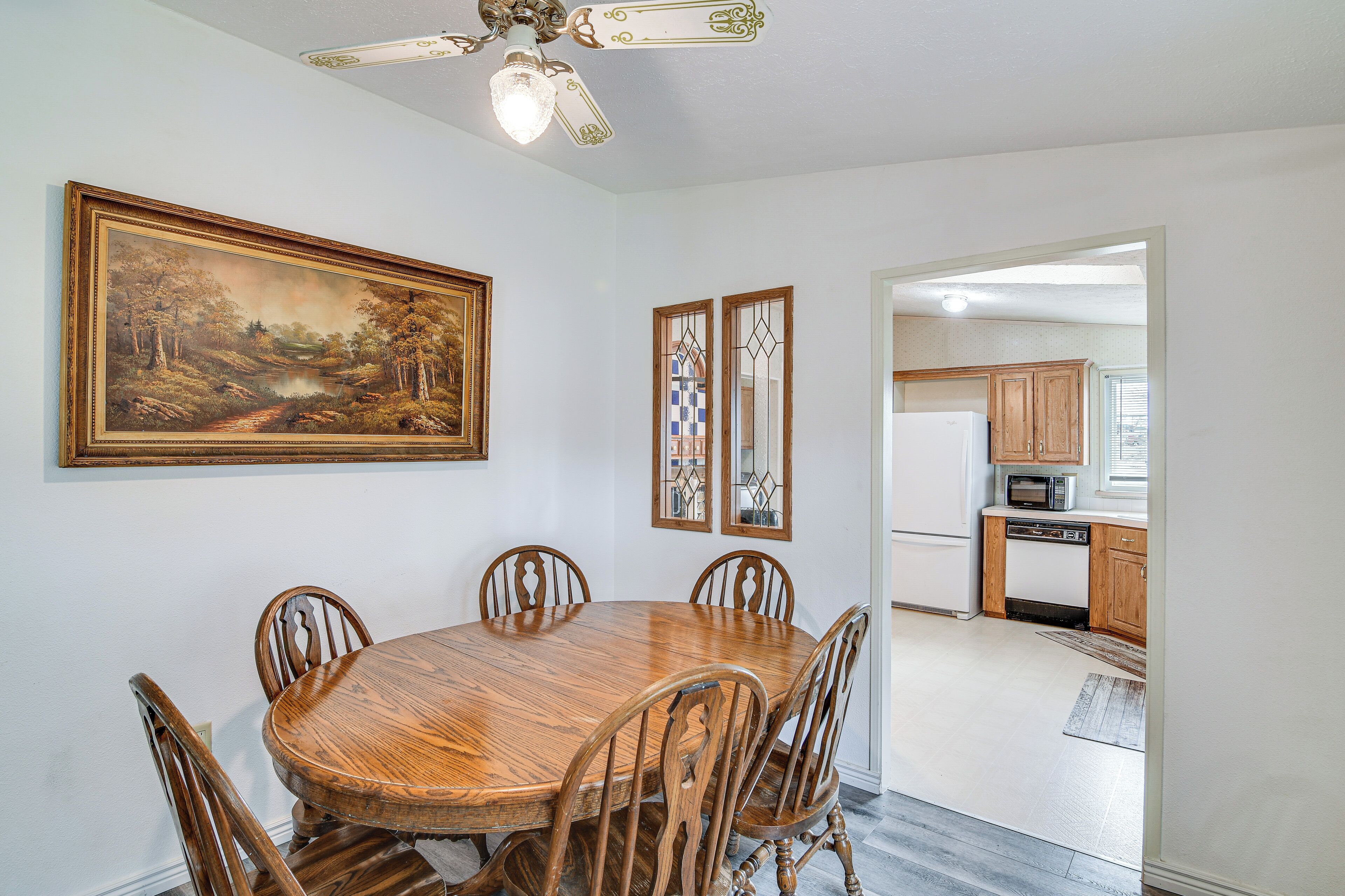 A dining room with a wooden table and chairs, a ceiling fan, and a painting on the wall.