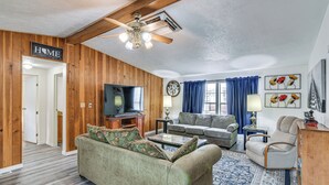 Una sala de estar con pared de madera, ventilador de techo, sofá, mesa de centro, televisor y ventana azul con cortinas.