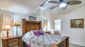 Un dormitorio con cabecera de madera, una cama con colcha estampada, dos mesitas de noche con lámparas, un ventilador de techo y un cuadro enmarcado en la pared.