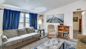 Una sala de estar con sofá, sillón y estantería, una ventana grande con cortinas y un cuadro en la pared.