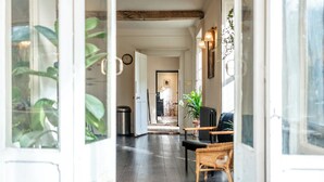 A hallway with a dark wooden floor, wicker furniture, and a view into a room with a bed and a plant. - Stunning 8-Bedroom Home in Painswick (Painswick)