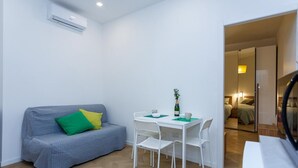 Ein kompakter Wohnraum mit einem grauen Sofa, einem kleinen weißen Tisch und einem weißen Stuhl. Eine Klimaanlage ist an der Wand angebracht.