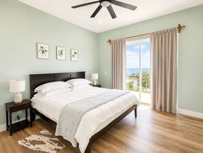 A bedroom with a bed, bedside lamps, a ceiling fan, a window with curtains, and a view of the outdoors. - Ocean Crest  6BR + Private Pool & Ocean Views (Camp Bay)