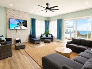 A modern living room with a flat-screen TV, wicker furniture, a ceiling fan, and a view of the ocean.